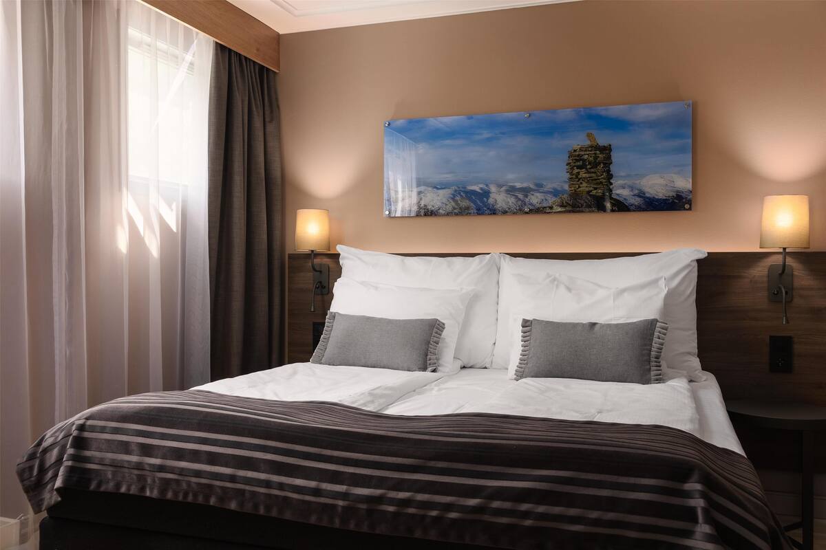 Ein Hotelzimmer mit Bett, zwei Nachttischlampen, einem Berglandschaftsbild und einem Fenster mit durchsichtigen Vorhängen.