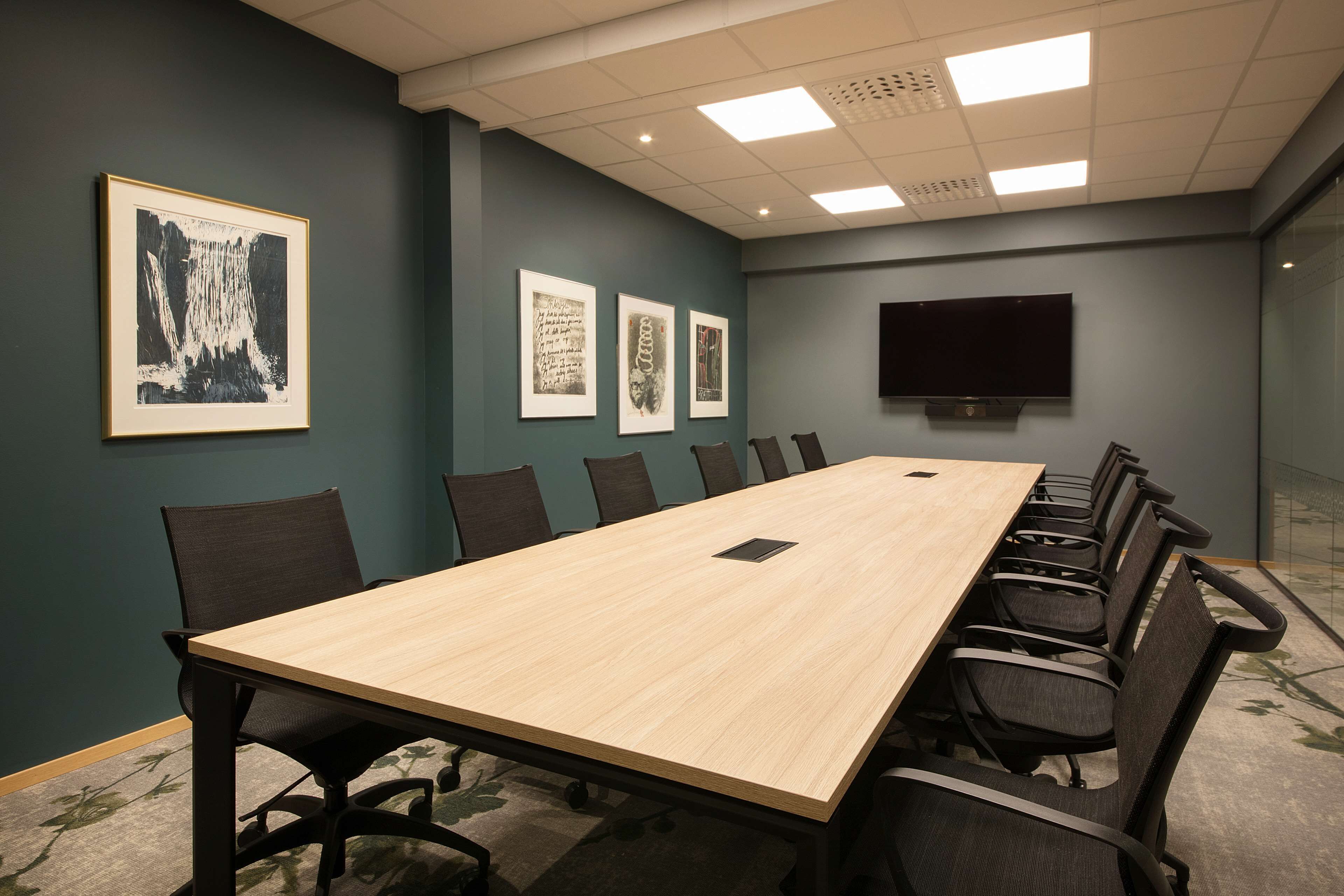 A conference room with a long table, chairs, framed artwork on the wall, and a flat-screen TV.