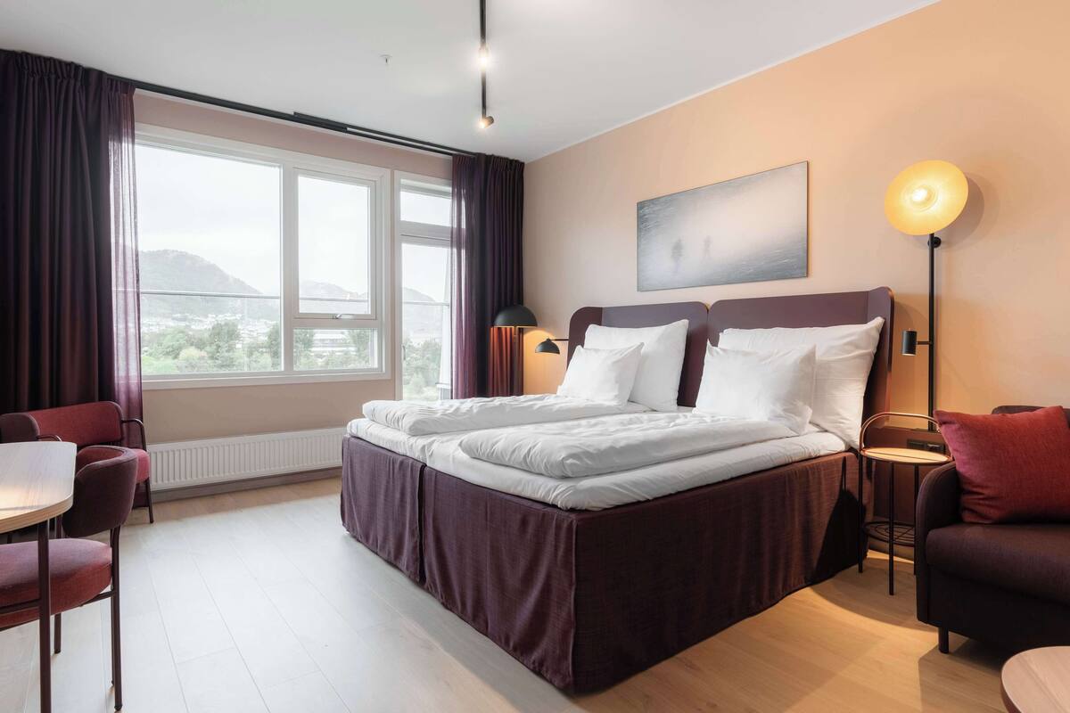 Ein Hotelzimmer mit einem großen Bett, zwei Nachttischen, einem Sessel und einem Tisch, sowie Blick ins Freie.