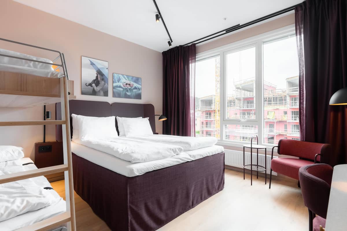 Ein Hotelzimmer mit Etagenbett, einem Einzelbett, einem roten Sessel, einem kleinen Tisch und einem großen Fenster mit Blick auf die Stadt.