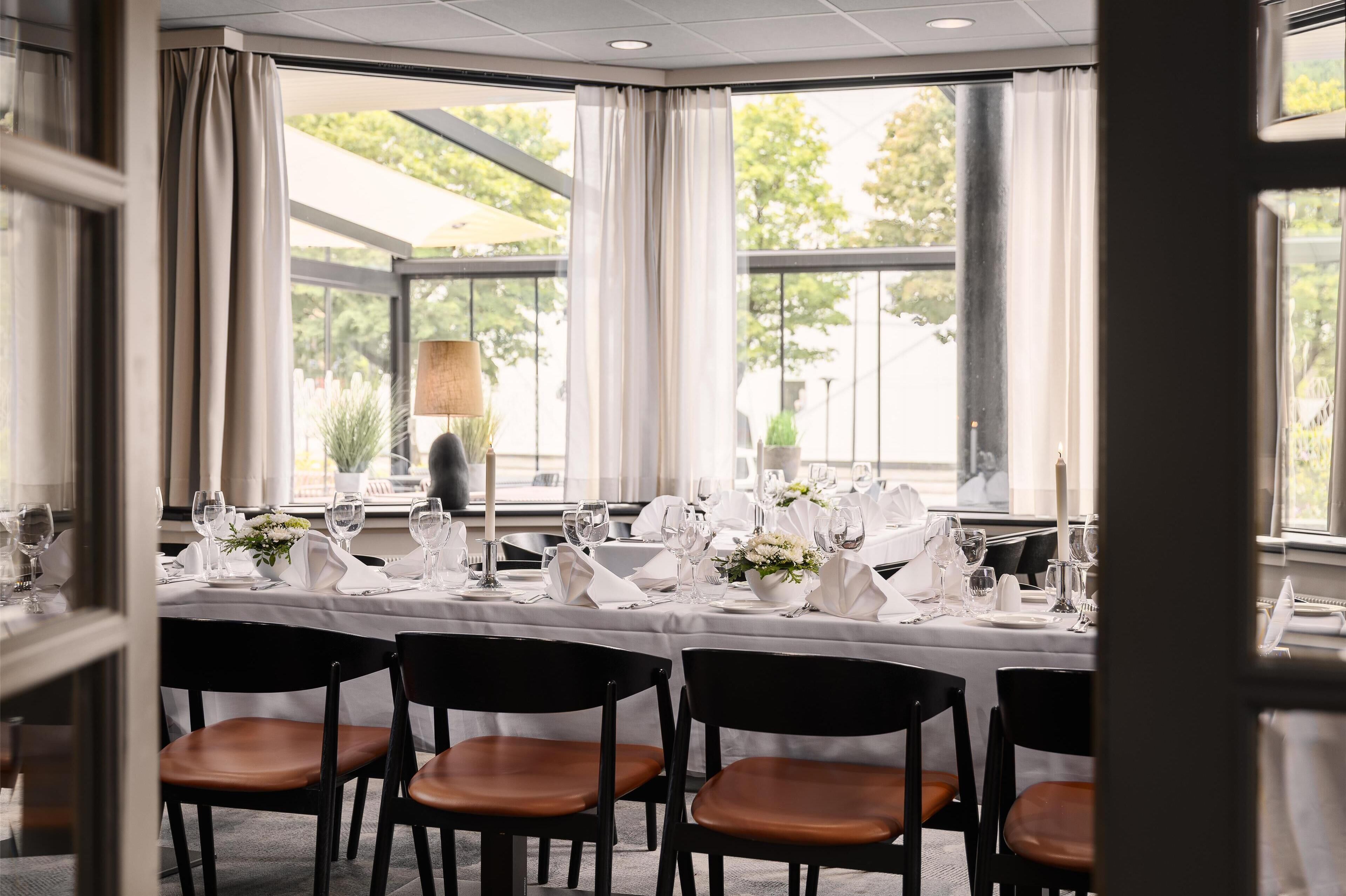 Un comedor con una mesa larga dispuesta para una comida, rodeada de sillas y grandes ventanales con cortinas.