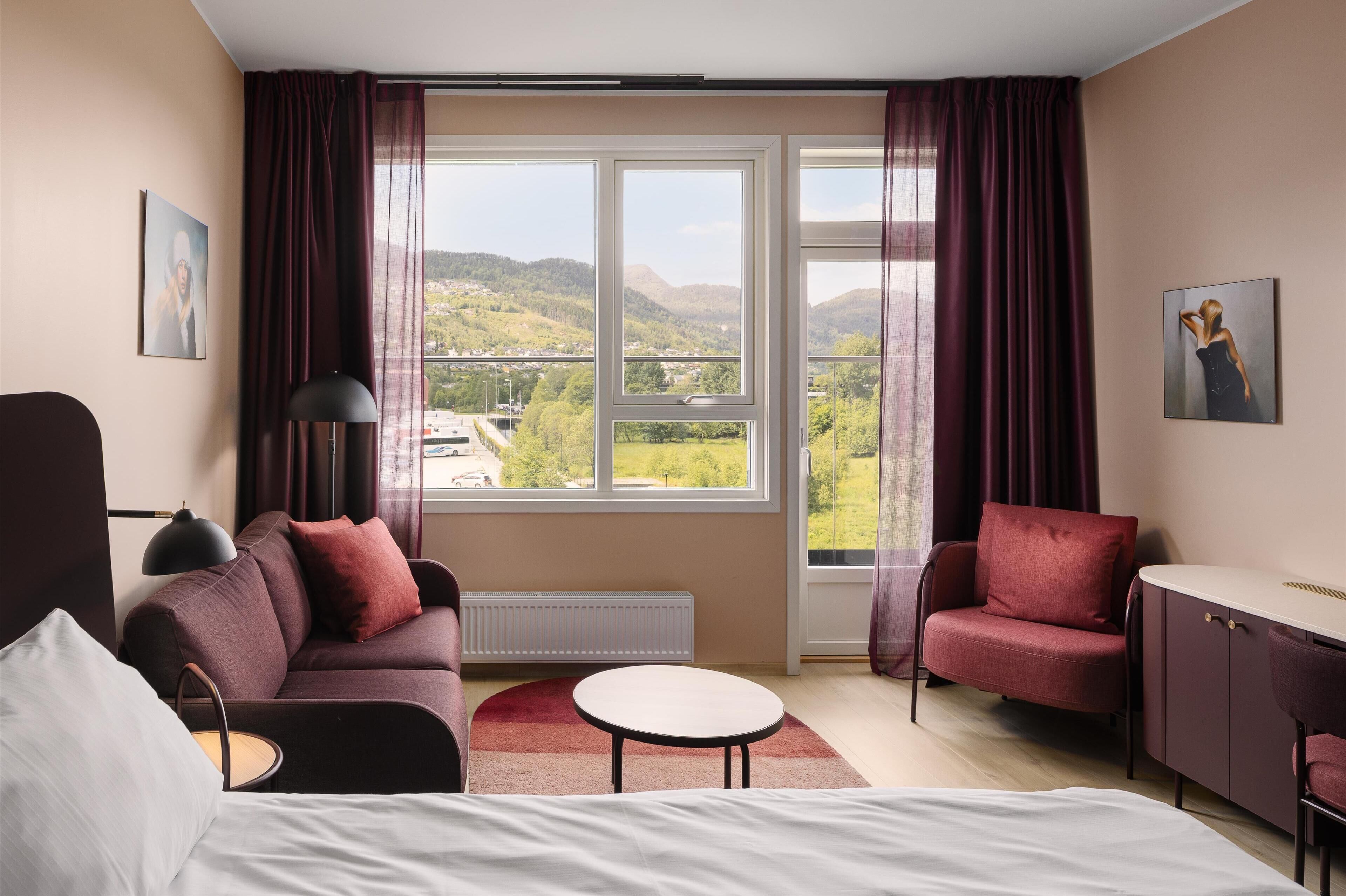 A modern hotel room with a large window, a sofa, a desk, and a bed.