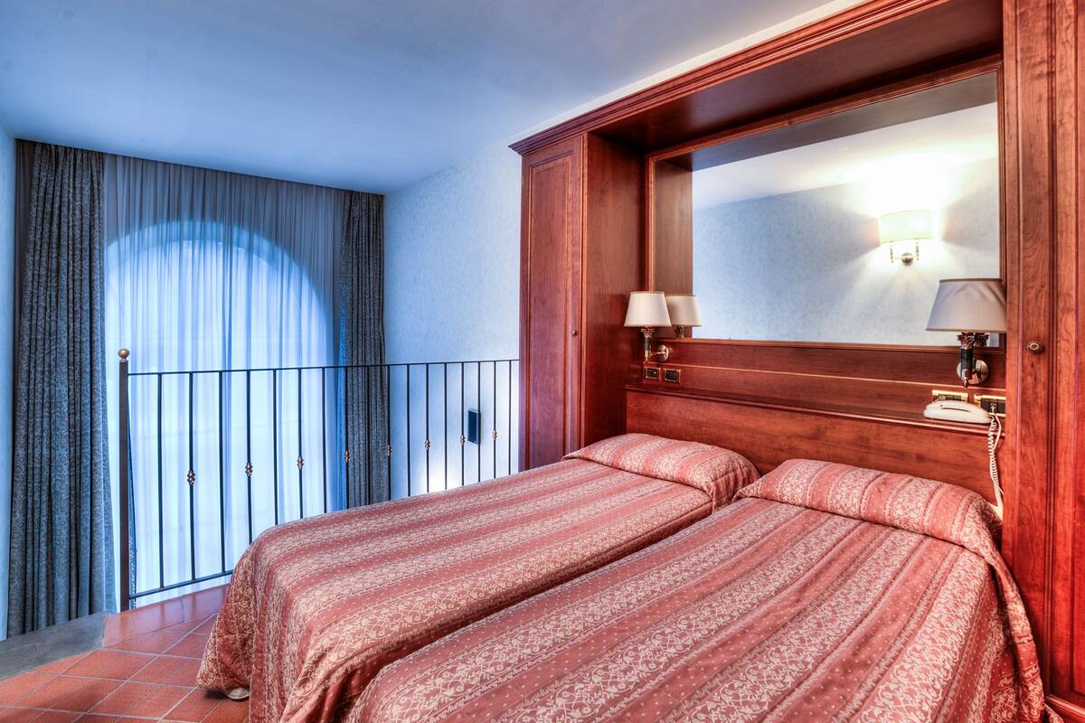 Una habitación de hotel con una cama grande, cabecera de madera, espejo y balcón con cortinas.