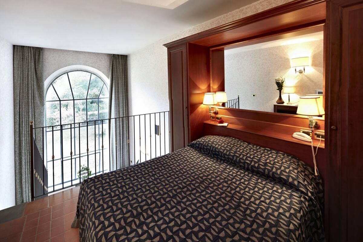 Habitación de hotel con una cama grande, un balcón con barandilla, un espejo amplio y dos lámparas de noche.