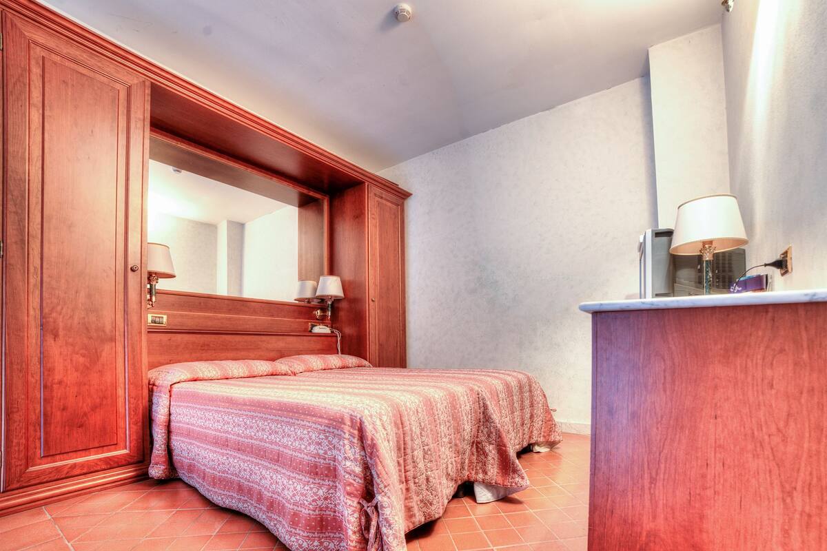 Un dormitorio con un armario de madera, una cama con colcha estampada, una mesita de noche con lámpara y un espejo sobre la cama.