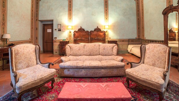 A spacious room with a large bed, two armchairs, a red ottoman, and a chandelier. - Hotel Villa Stanley (Sesto Fiorentino)