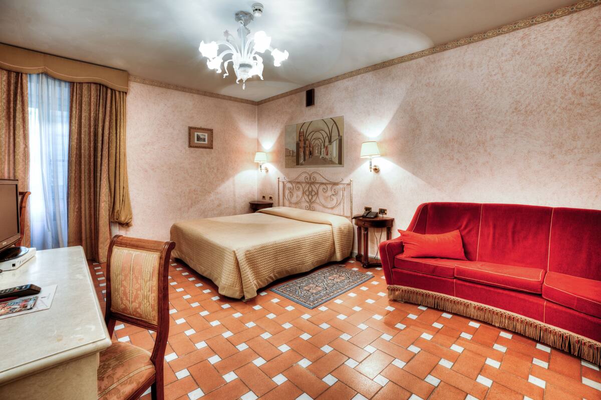 Una habitación de hotel con una cama, un sofá rojo, un escritorio con una computadora y un candelabro.