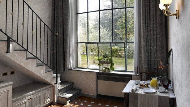 A room with a large arched window, a staircase with black railings, a small table with a lamp, and a radiator. - Hotel Villa Stanley (Sesto Fiorentino)