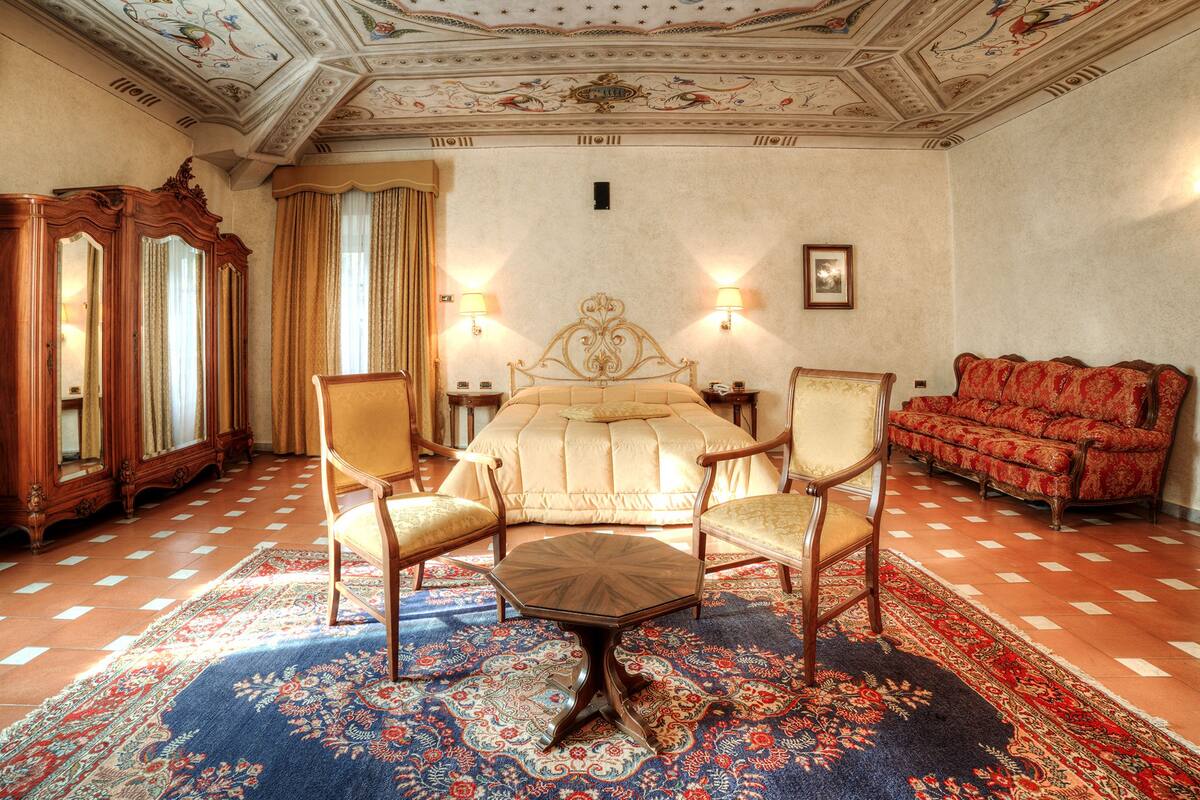 Un dormitorio lujoso con una cama grande, dos sillones, un sofá rojo, un techo ornamentado y una alfombra estampada.