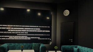 A modern lounge area with teal sofas and ottomans, a white table, and a wall with a clock and text. - Colour Hotel (Frankfurt)