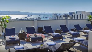 A rooftop terrace with lounge chairs and tables, offering a view of a harbor with ships and buildings in the background. - The Eliott Hotel (Gibraltar)