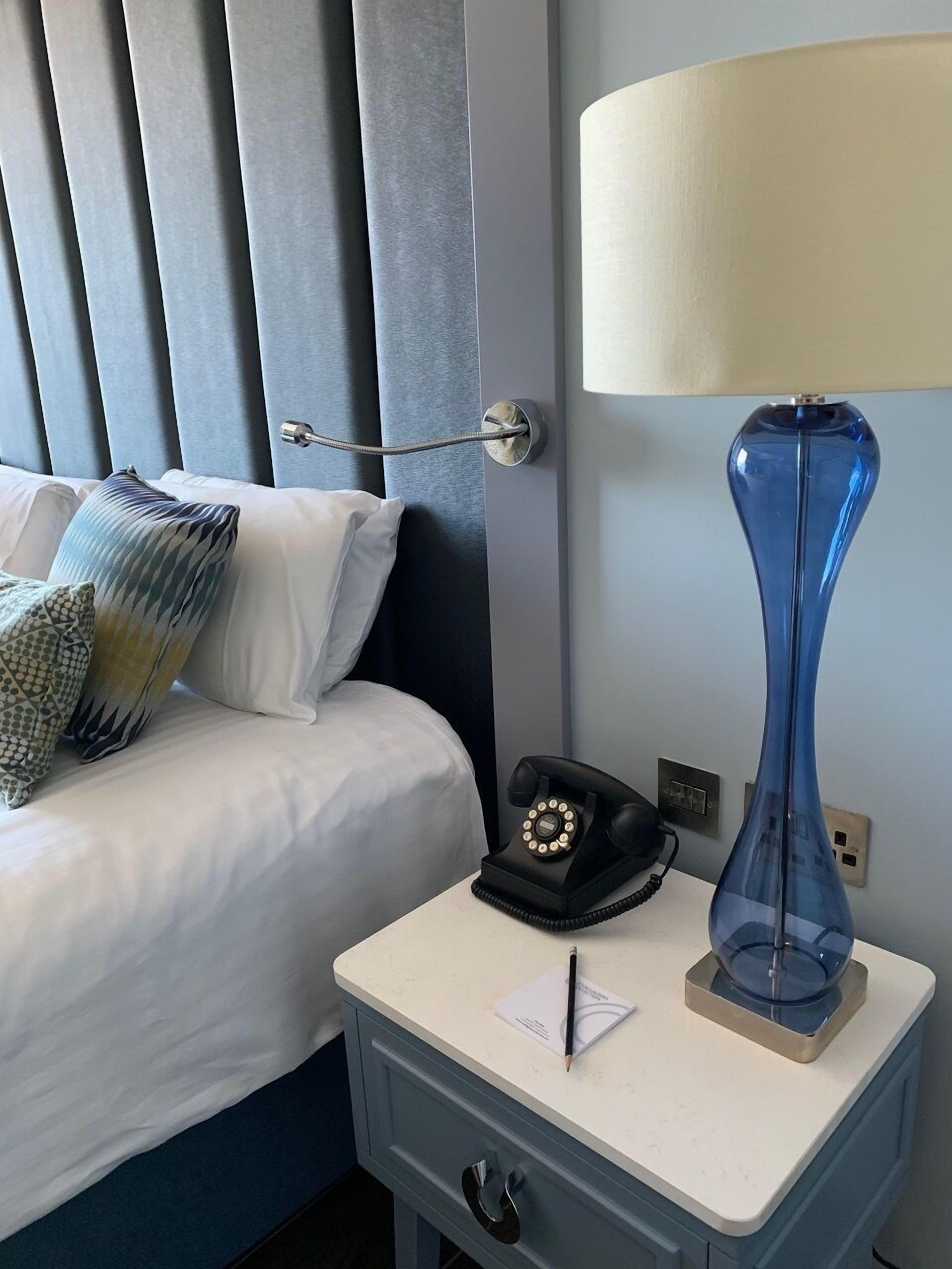 A hotel room with a bed, a bedside table with a lamp, a telephone, and a notepad.