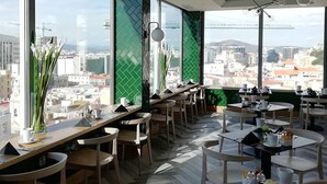 A modern cafe with large windows offering a city view, wooden tables and chairs, and a unique ceiling light fixture. - The Eliott Hotel (Gibraltar)
