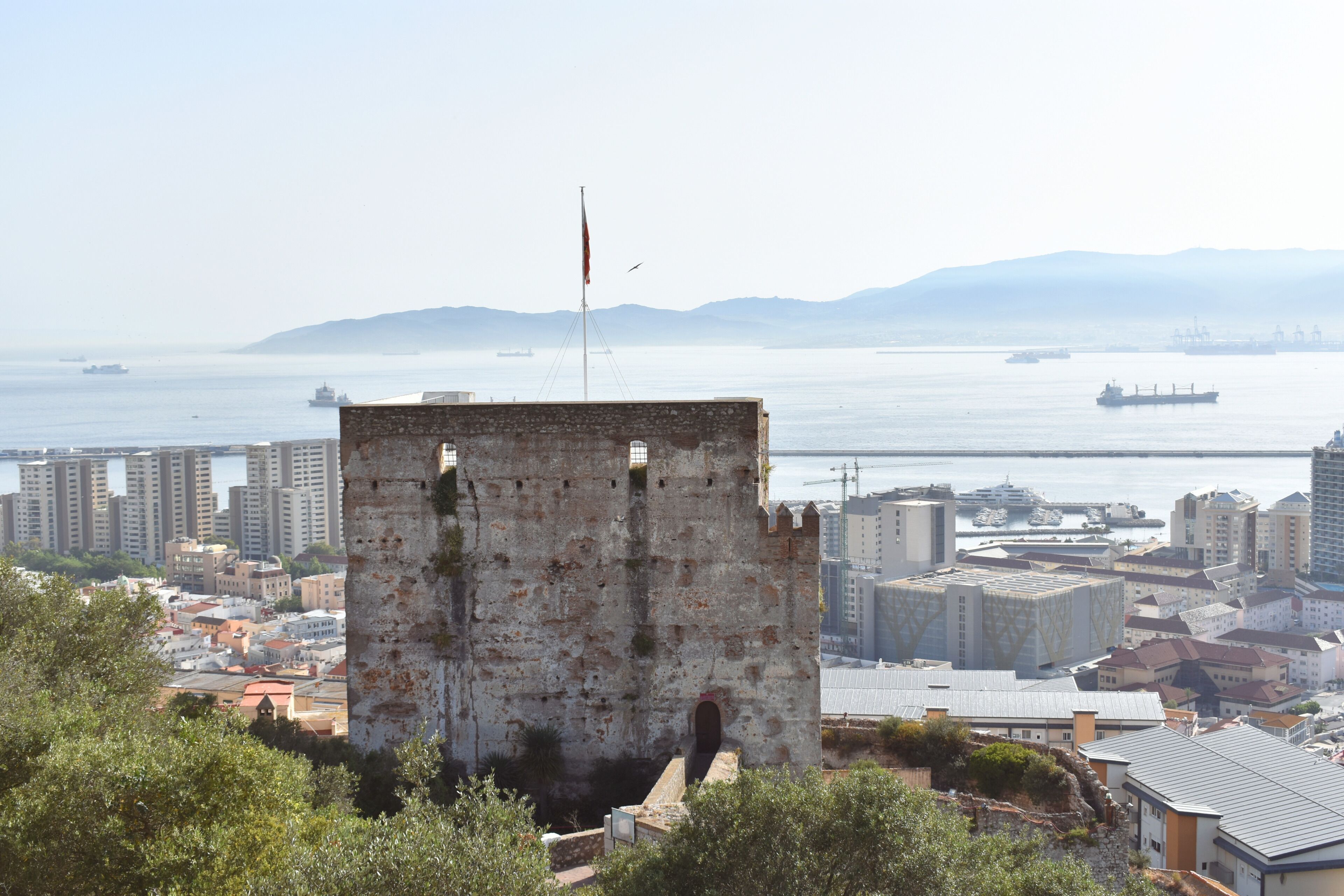 Uma torre histórica com uma bandeira, com vista para uma paisagem urbana costeira com edifícios modernos e um porto de iates.