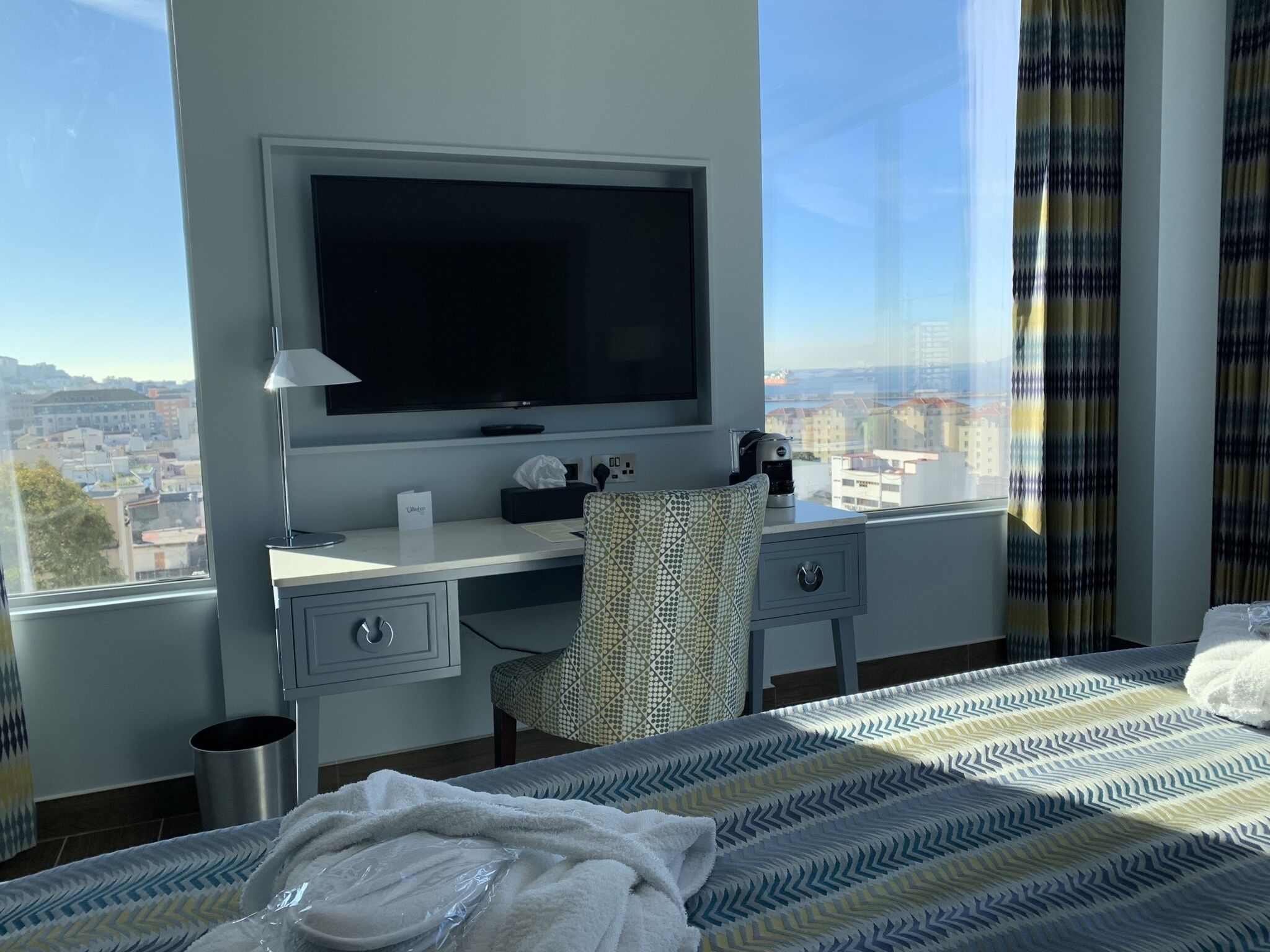 A hotel room with a large bed, a desk with a TV, a chair, and a view of the city.
