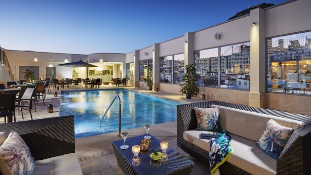 A rooftop pool area with seating and dining arrangements, surrounded by large windows offering city views. - The Eliott Hotel (Gibraltar)