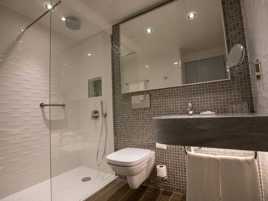 Un baño moderno con ducha envuelta en vidrio, inodoro blanco y un espejo grande sobre un lavabo con encimera oscura.
