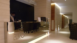 Balcão de recepção de hotel com design moderno, uma tela digital e uma área de estar com cadeiras.