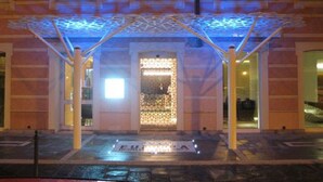 Entrada de edifício com marquise, porta decorativa e pequena placa iluminada.