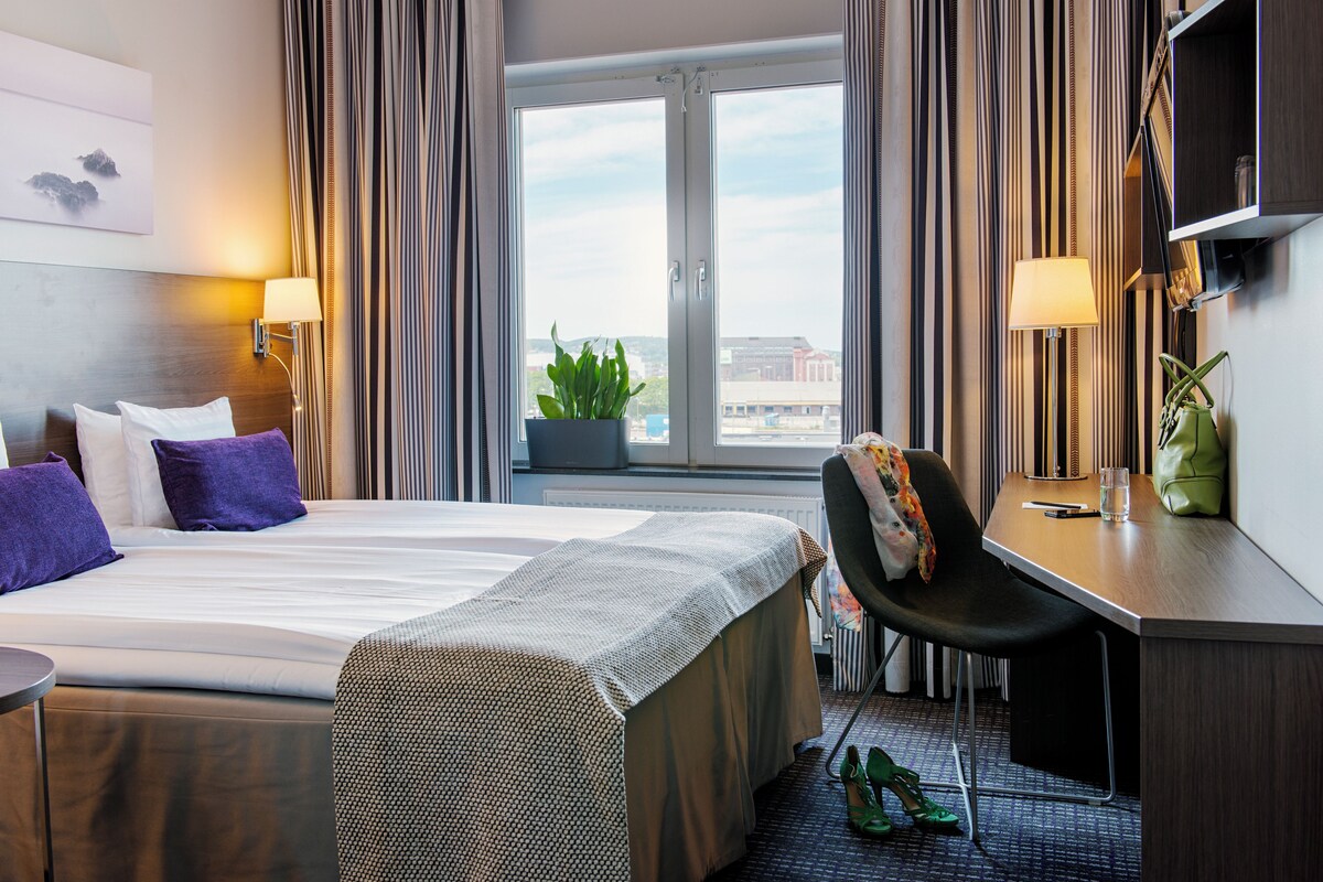 A hotel room with a bed, a desk, a chair, a potted plant, and a view of the city through the window.