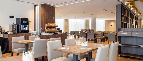 A modern hotel dining area with multiple tables and chairs, a kitchen area, and a reception desk.