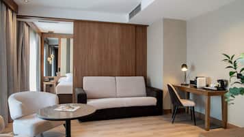 A modern hotel room with a wooden panel wall, a sofa, a round table, a desk with a lamp, and a plant.