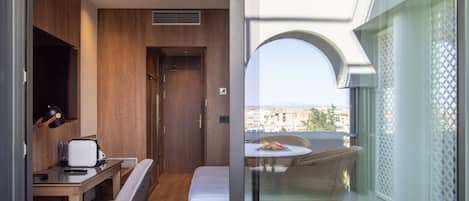 Habitación compacta con escritorio de madera, una silla, una cama y un balcón con mobiliario de mimbre.
