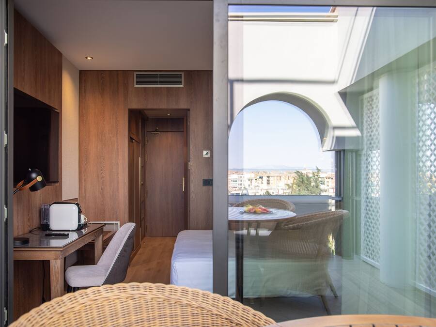 A compact room with a wooden desk, a chair, a bed, and a balcony with wicker furniture.