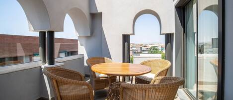 Un balcon agrémenté de meubles en osier, d’une table ronde et de grandes fenêtres donnant sur des immeubles.