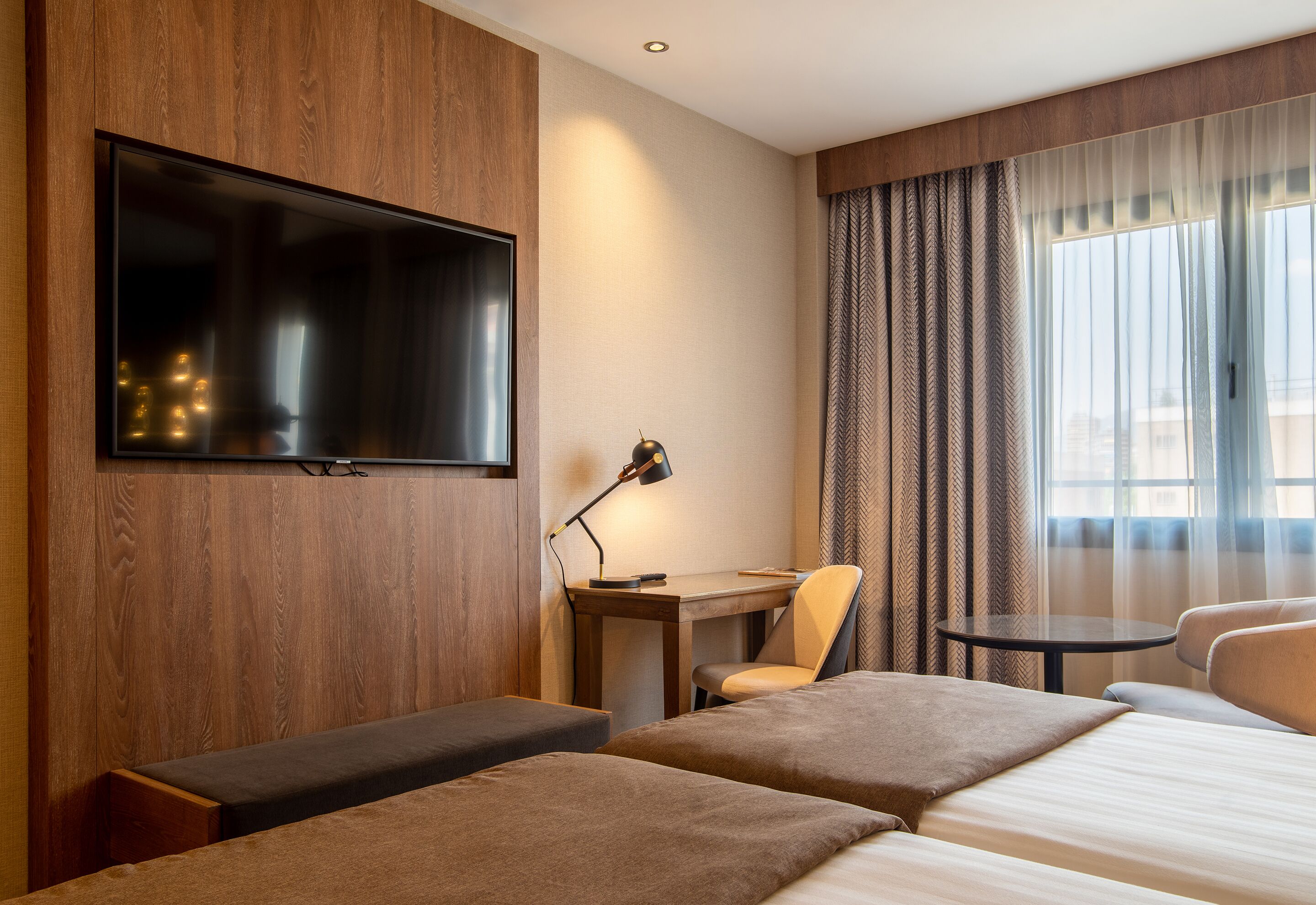 A hotel room with a large flat-screen TV mounted on the wall, a desk with a lamp, a chair, a small round table, and two beds with headboards.