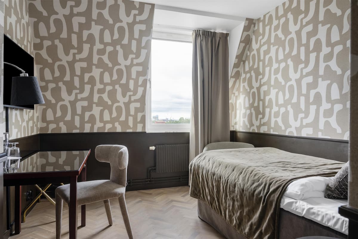 A hotel room with a bed, a desk, and a chair. There is a window with curtains, and the walls have a patterned wallpaper.