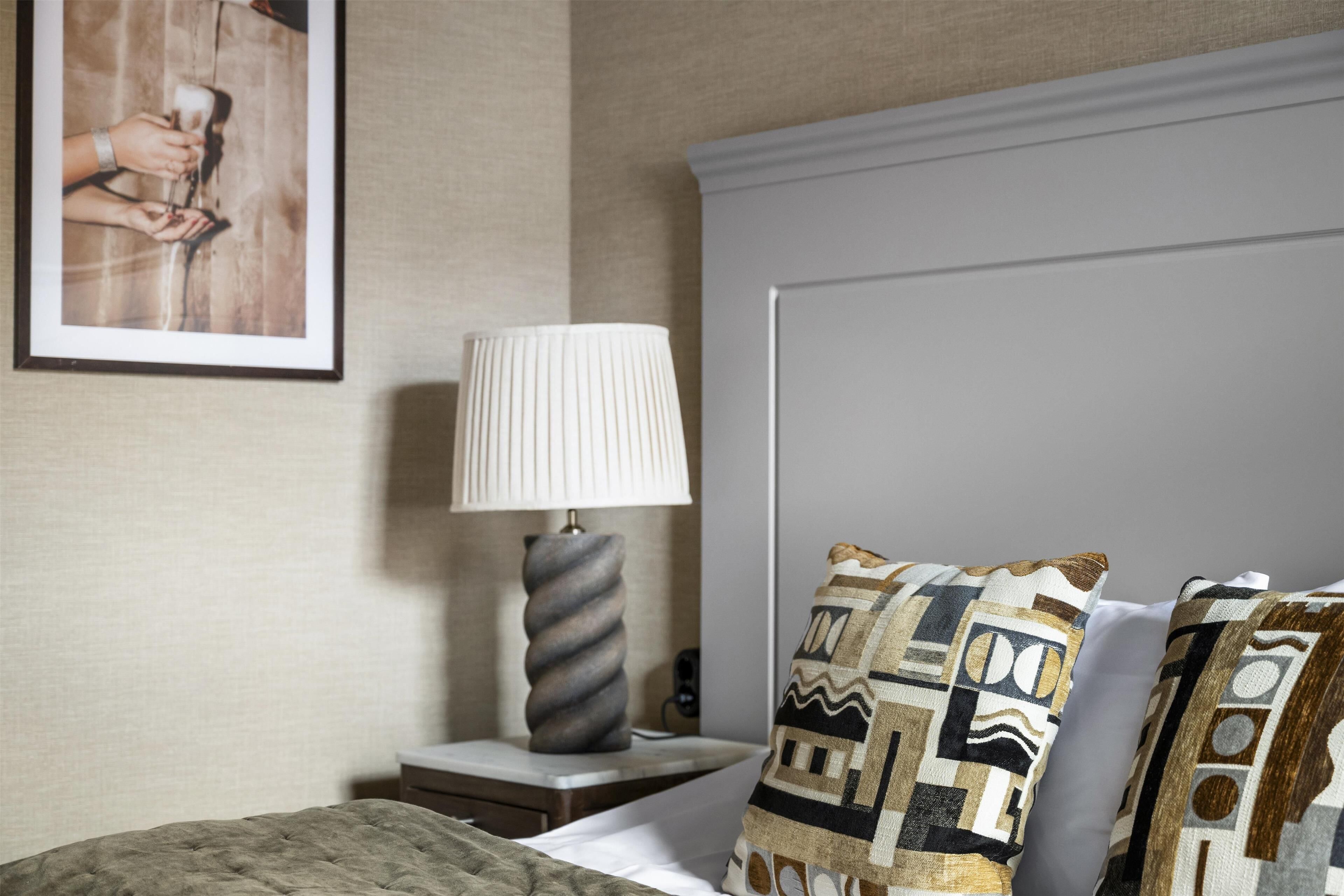 A neatly arranged hotel room with a bed, a bedside lamp, a framed picture on the wall, and patterned pillows.