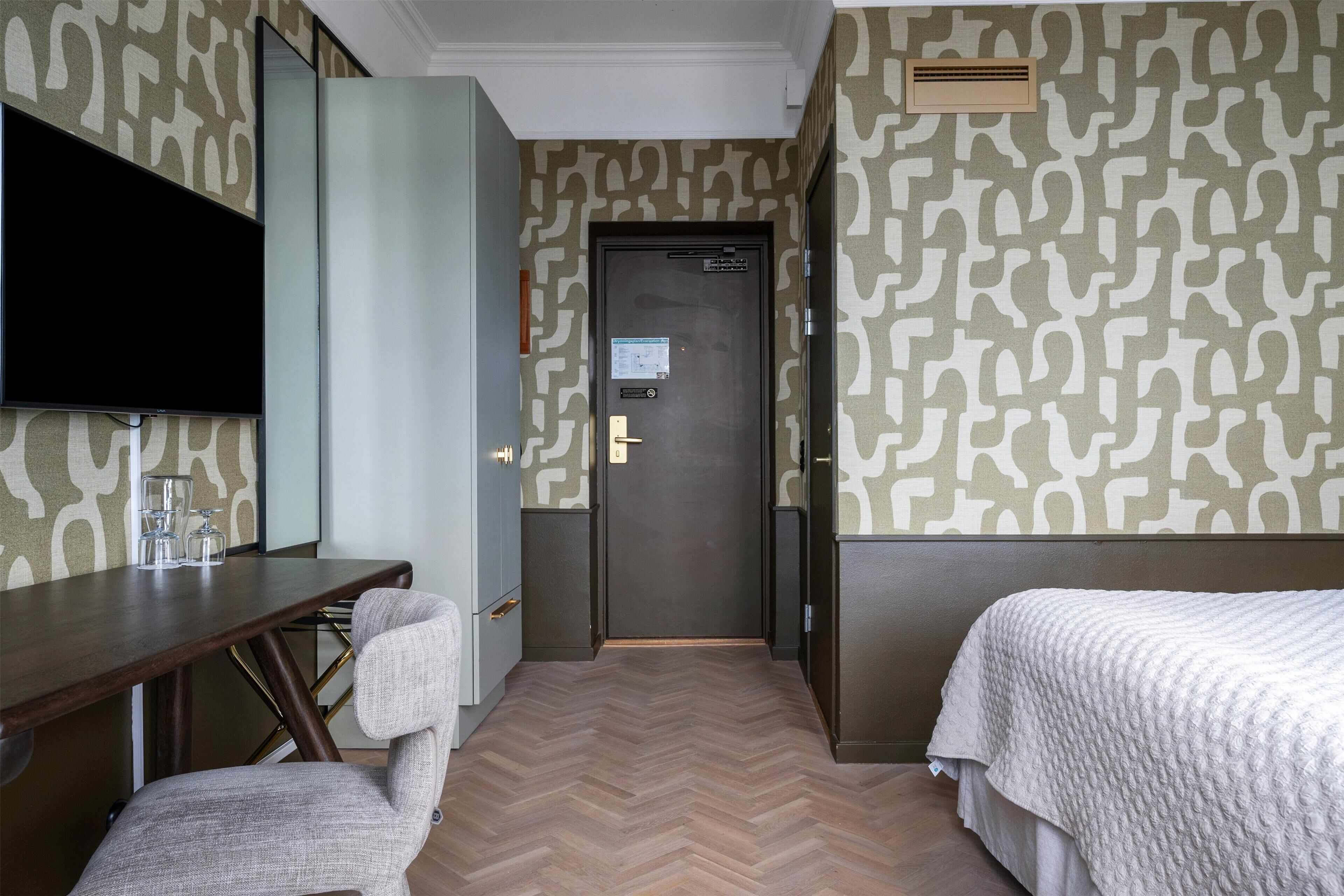 Une chambre d’hôtel avec un parquet en bois disposé en motif «chevron», un téléviseur à écran plat fixé au mur, un bureau avec une chaise et deux lits avec du linge de lit blanc.