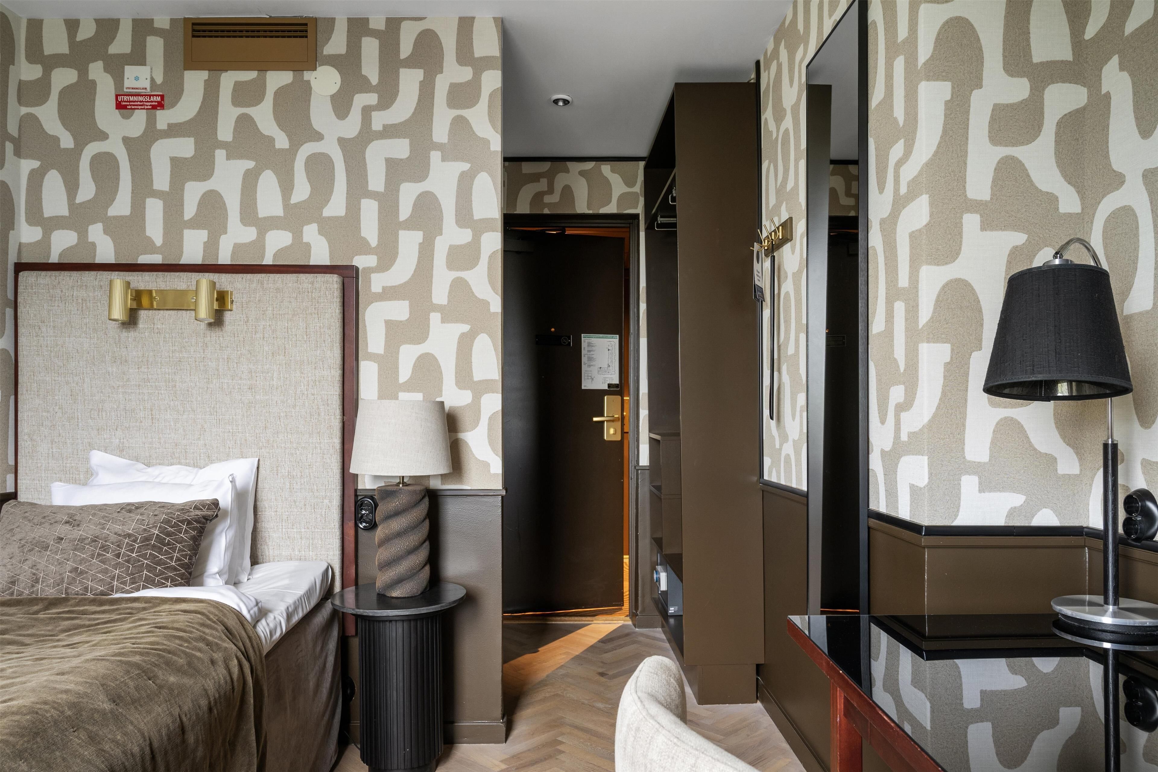 Una habitación de hotel con cama, mesita de noche, lámpara y espejo. Se ve una puerta que da a otra habitación.