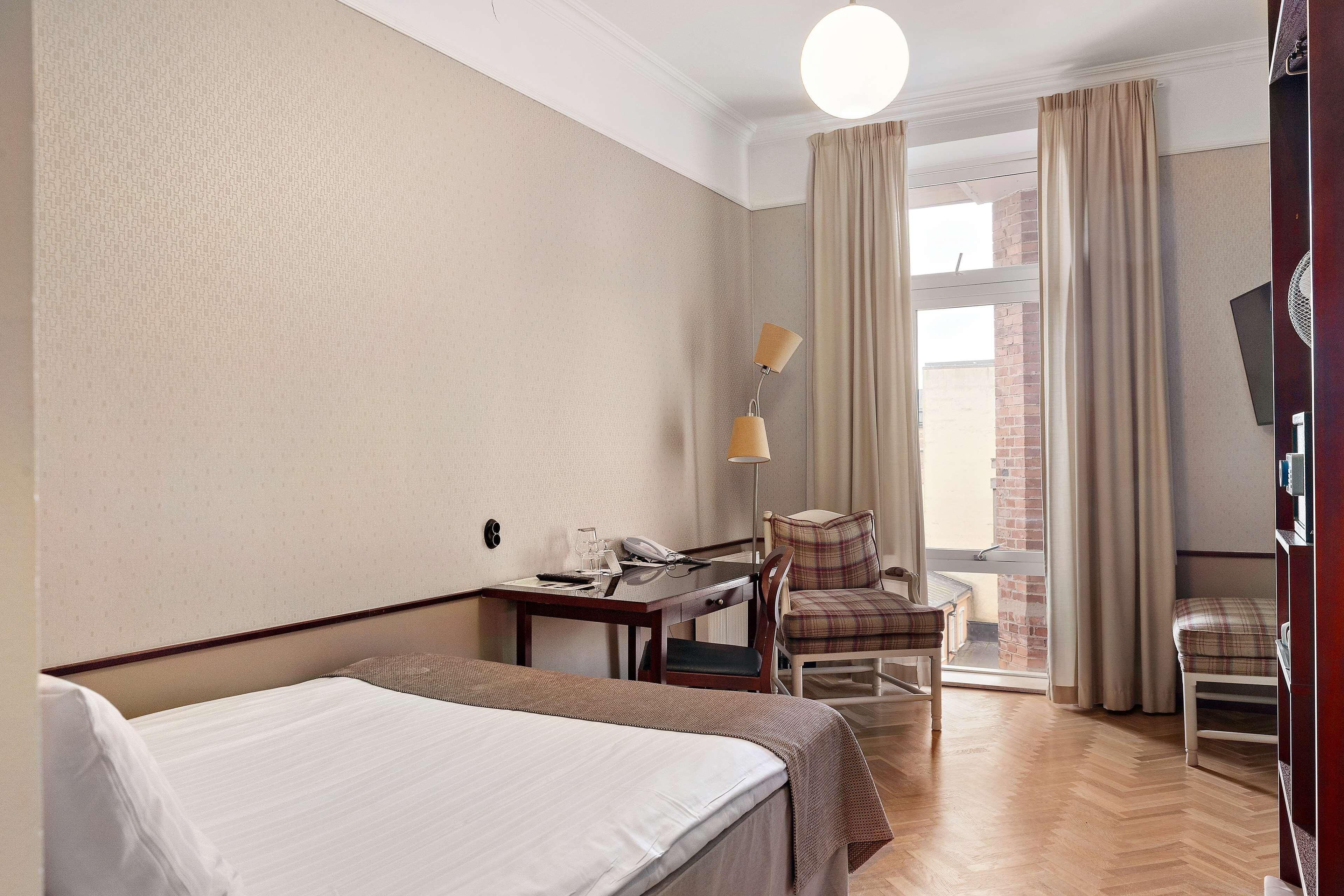 A hotel room with a bed, a desk with a telephone, a chair, a lamp, and a view of a brick building through the window.