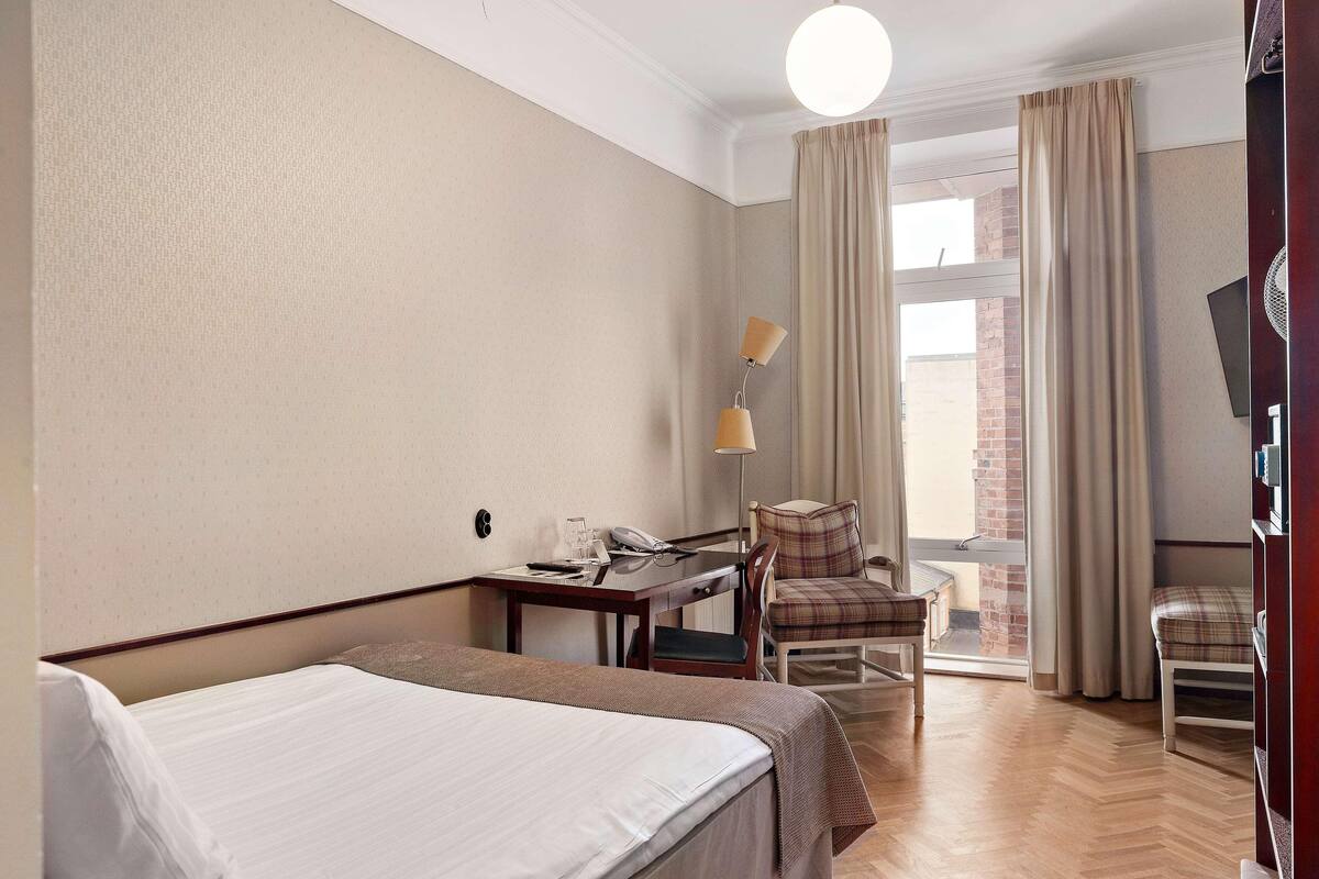 A hotel room with a bed, a desk with a telephone, a chair, a lamp, and a view of a brick building through the window.
