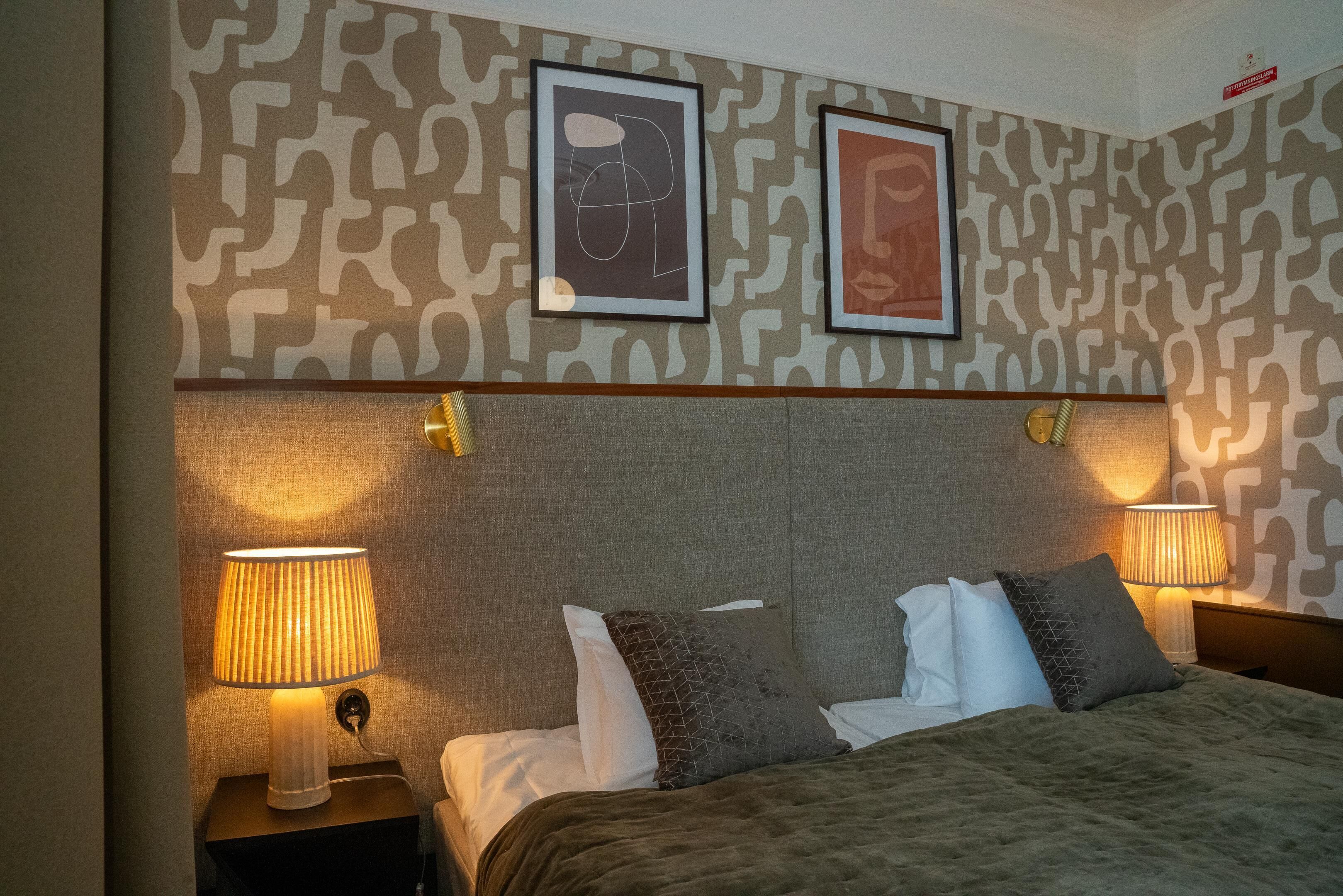 A hotel room with a patterned wallpaper, a large bed with multiple pillows, two bedside lamps, and two framed artworks on the wall.