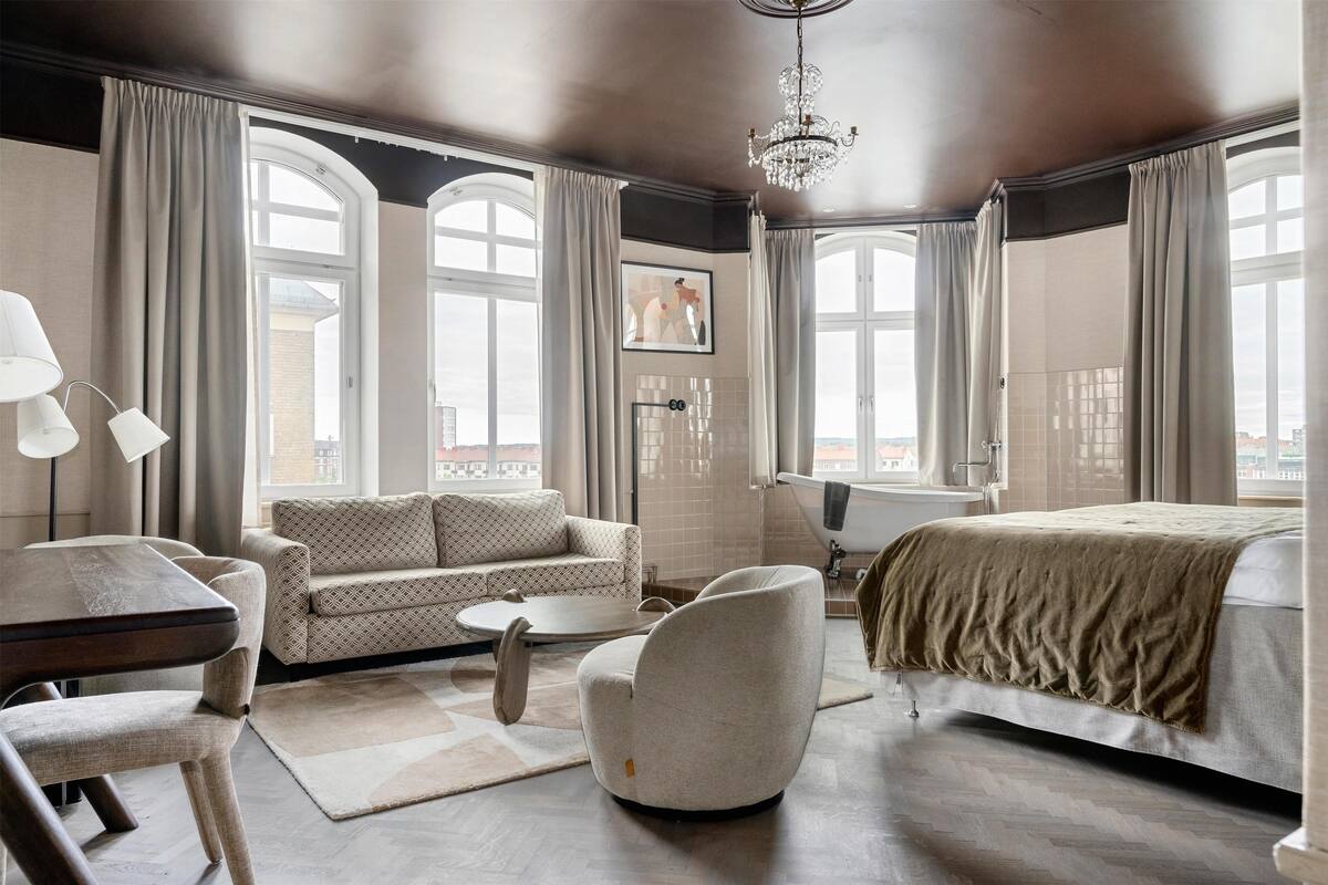 A modern hotel room with a large bed, a sofa, a dining table, and a chair. There are large windows with curtains, a chandelier, and a painting on the wall.
