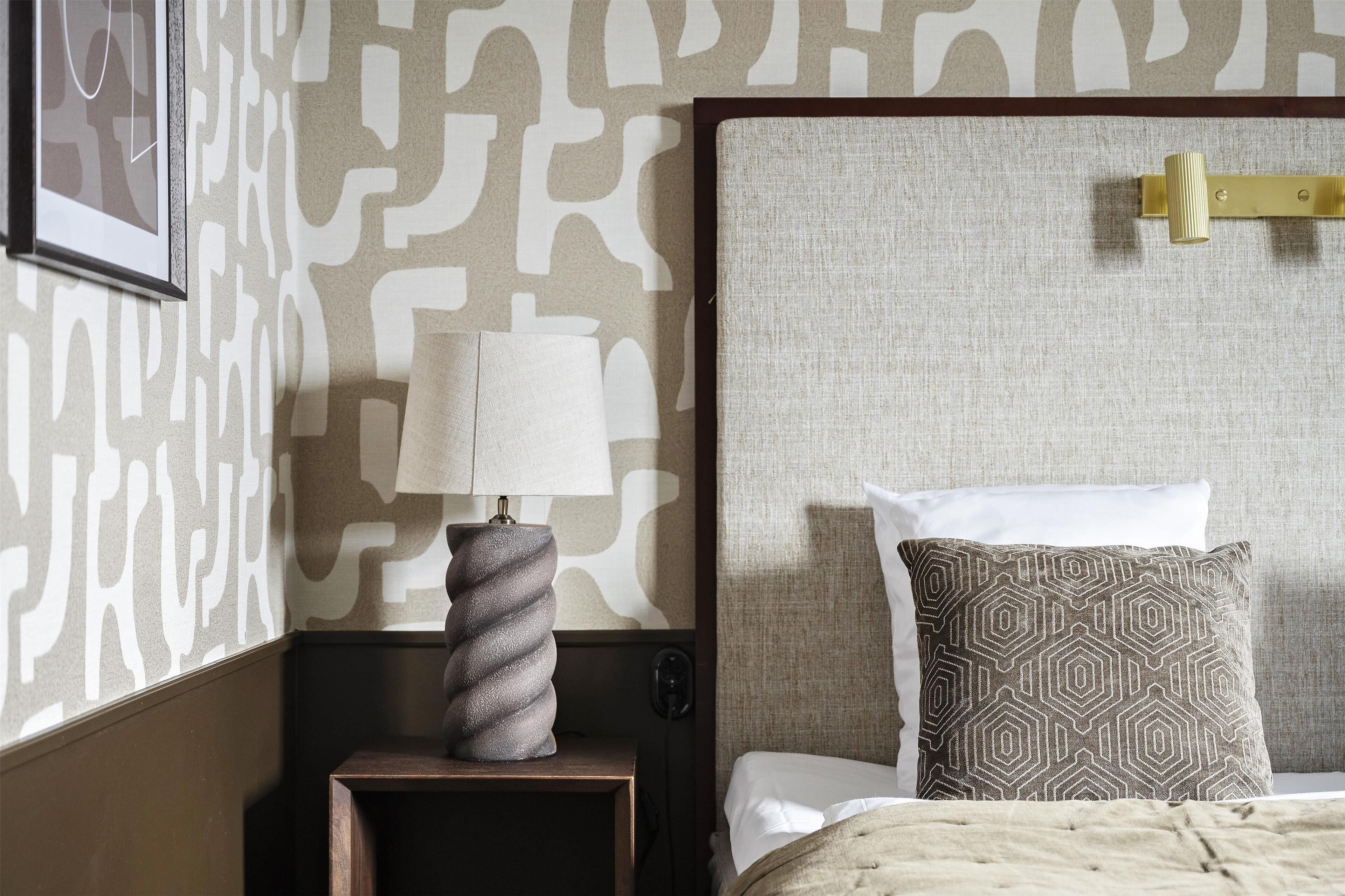 A neatly made bed with a patterned pillow, a bedside lamp on a wooden nightstand, and a wall with a geometric pattern.