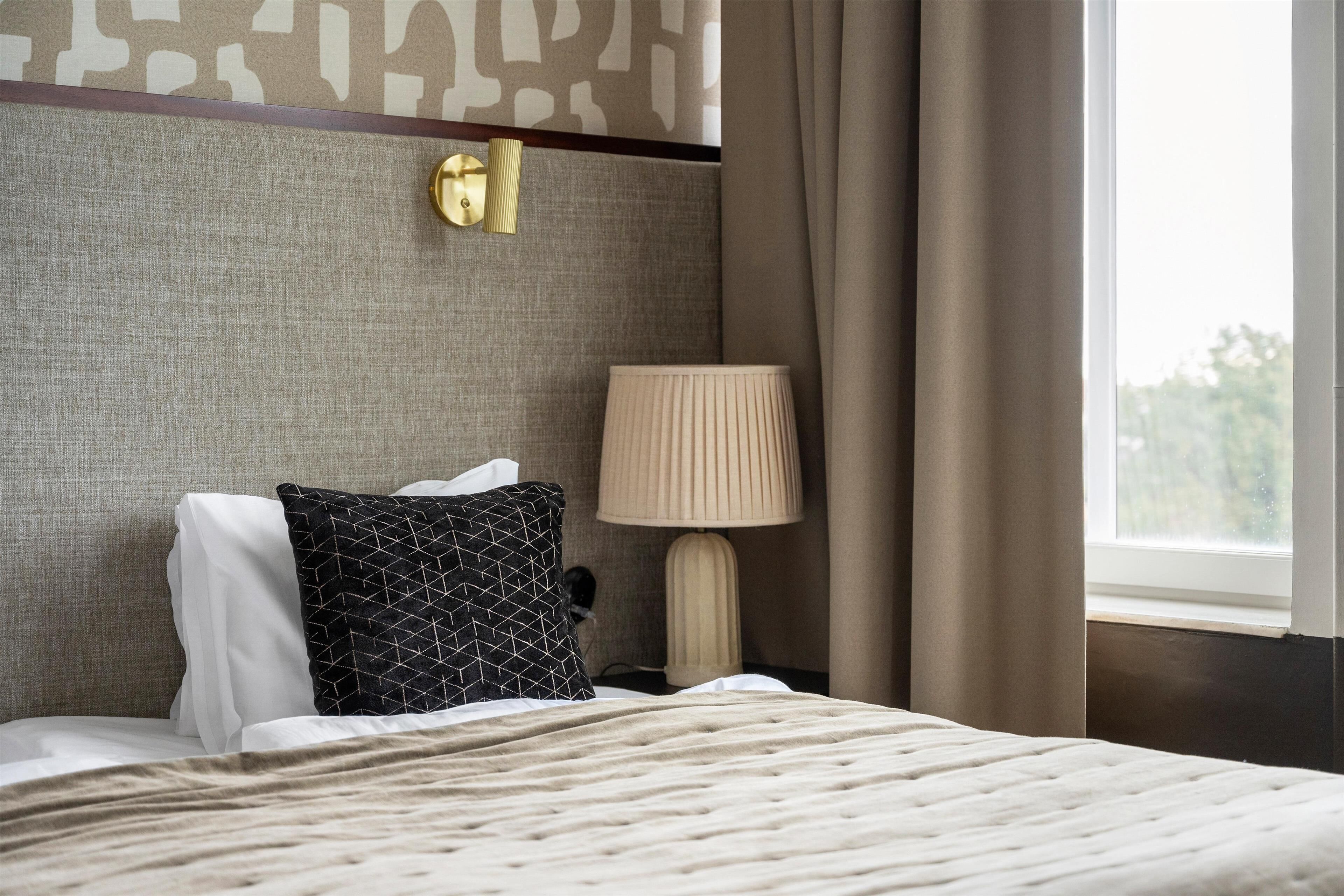 Una cama bien hecha con un edredón texturizado, una almohada de diseño blanco y negro y una lámpara color beige, junto con una lámpara fijada a la pared.