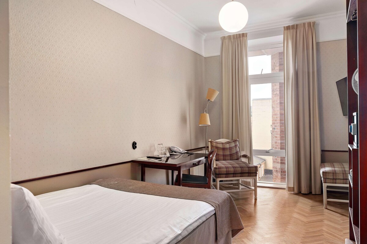 A hotel room with a bed, a desk with a telephone, a chair, a lamp, and a view of a brick building through the window.