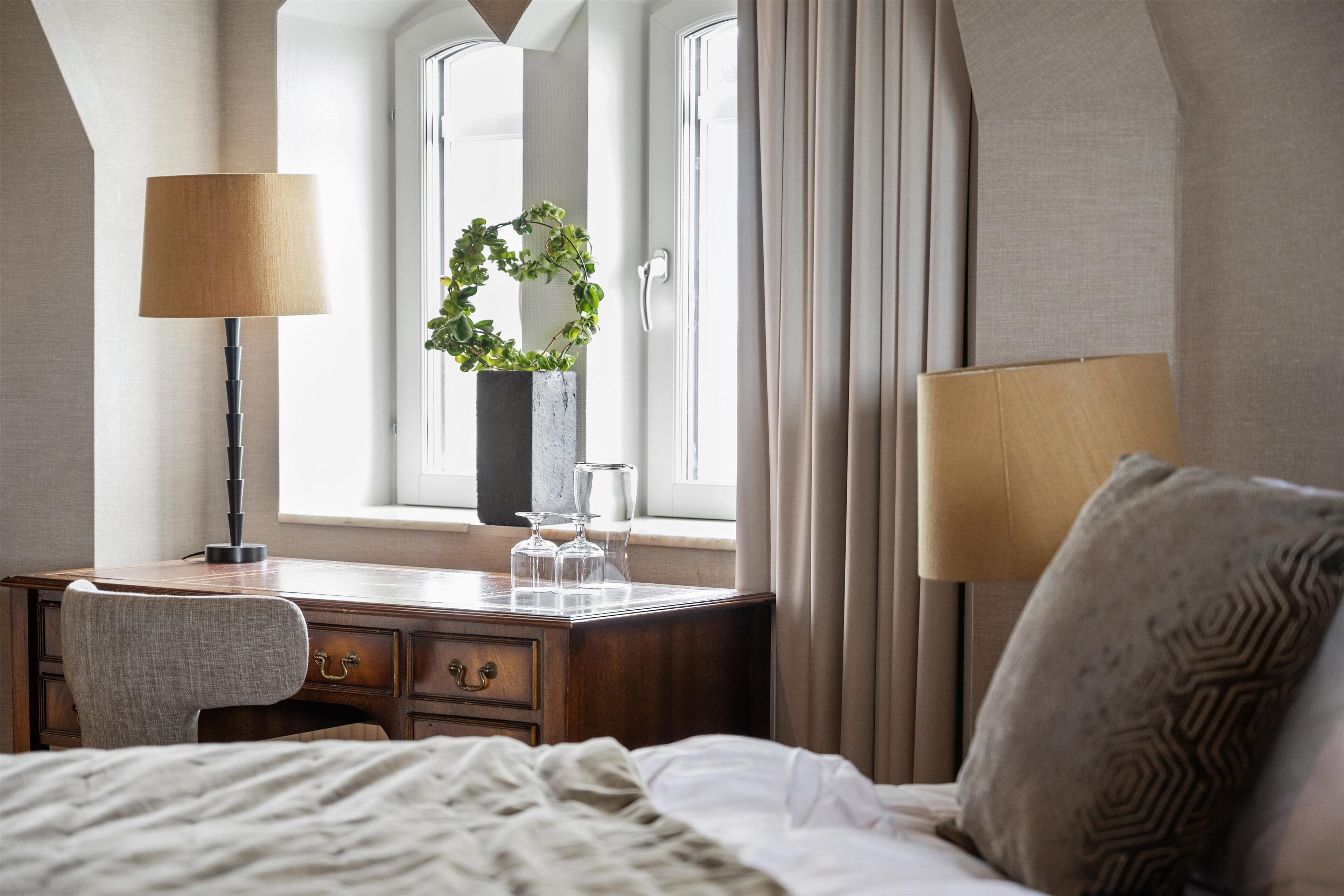A hotel room with a wooden desk, two lamps, a bed, a window with curtains, and a decorative wreath.