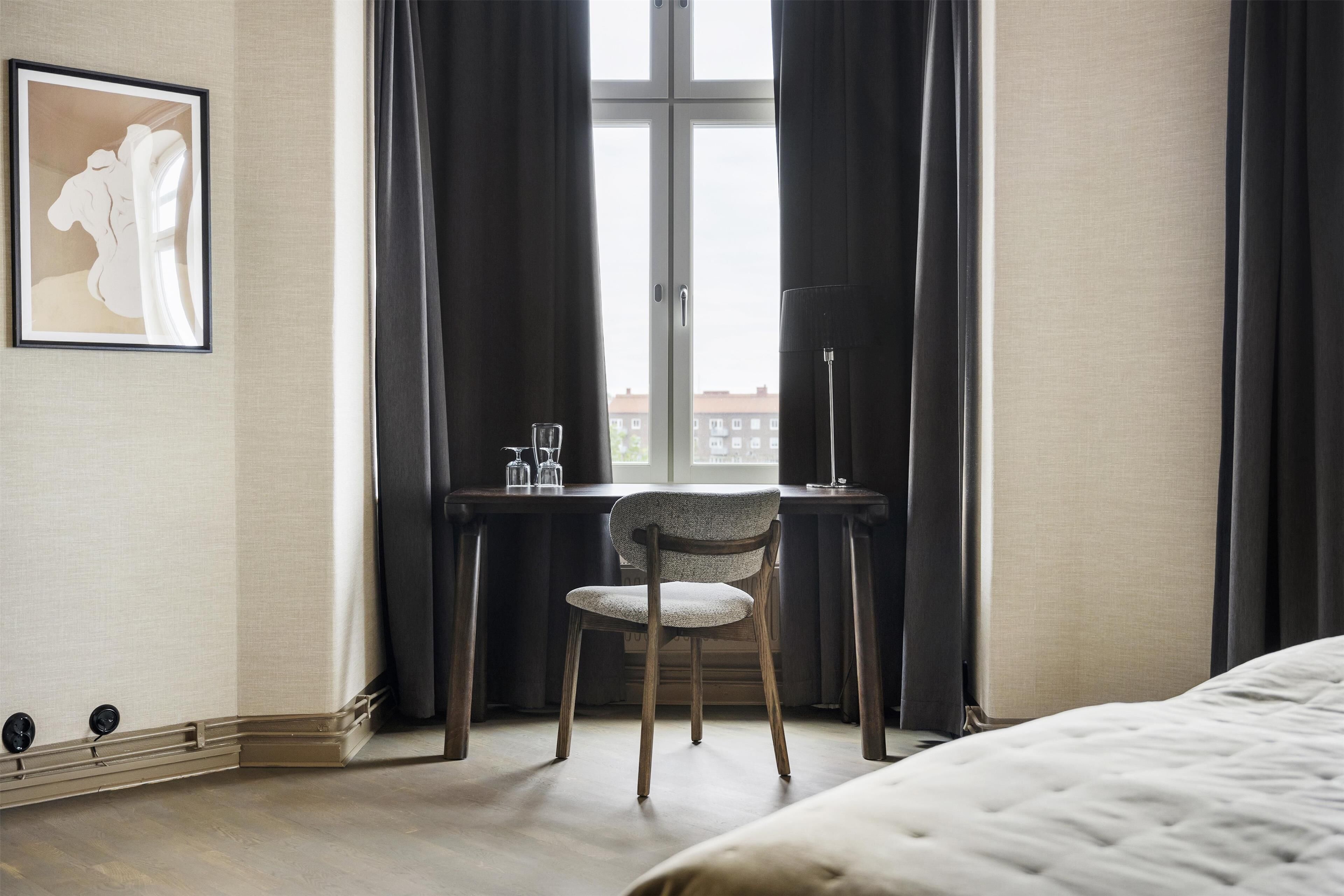A hotel room with a bed, a desk with a chair, a window with curtains, and a framed picture on the wall.