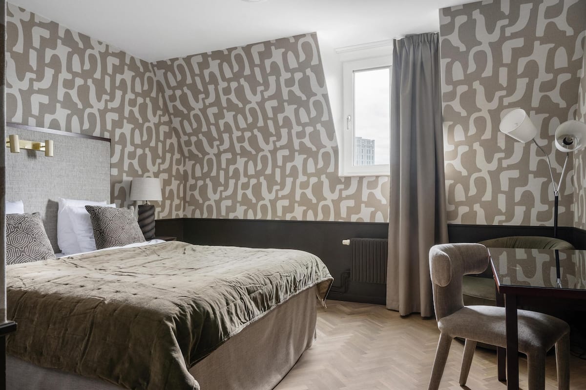 A hotel room with a large bed, a desk with a chair, a lamp, and patterned wallpaper.
