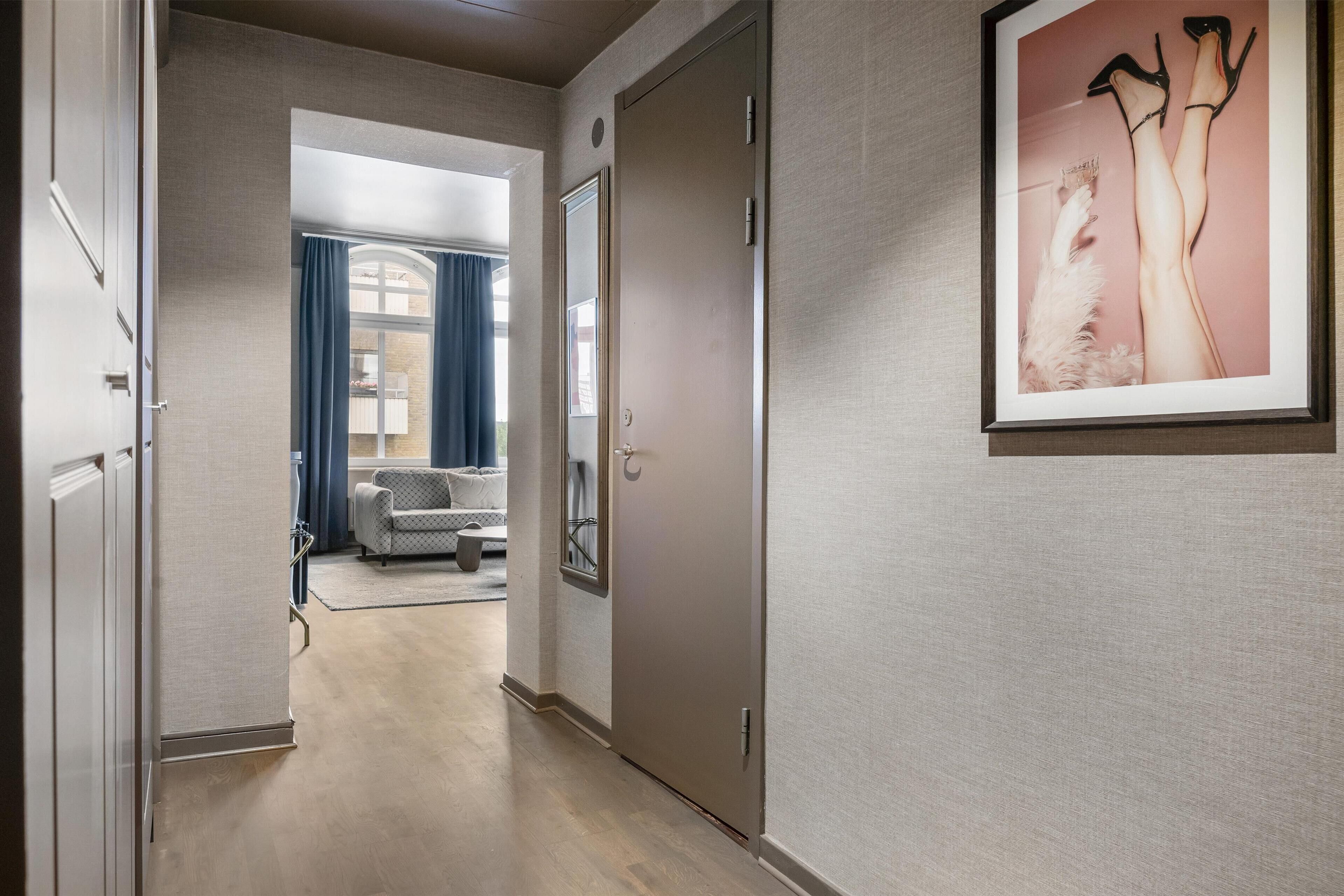 Un pasillo con un cuadro enmarcado de piernas con tacones altos, una puerta y una vista parcial de una sala de estar con sofá y cortinas.
