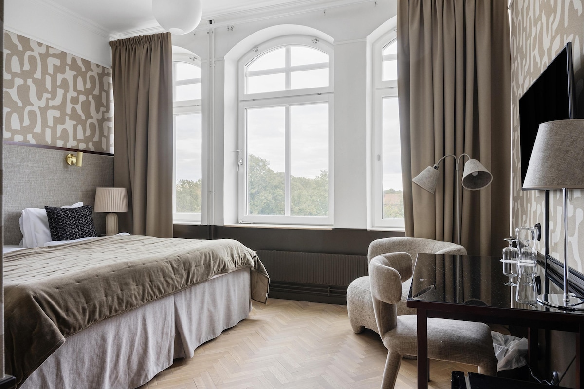 A hotel room with a large bed, a desk with a lamp, a chair, and a view of the outdoors through large windows.