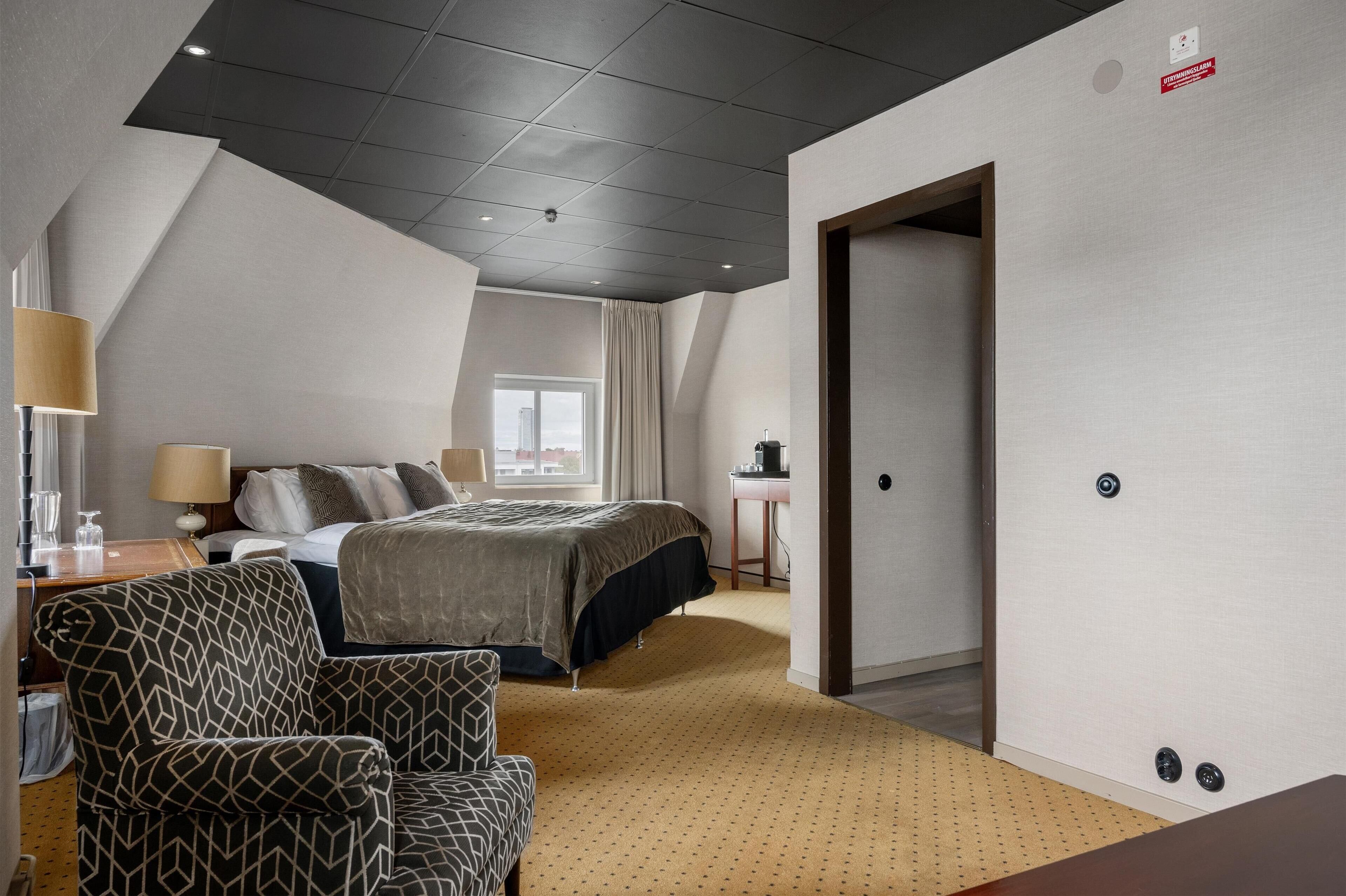 A hotel room with a bed, a desk, a chair, a lamp, and a window.