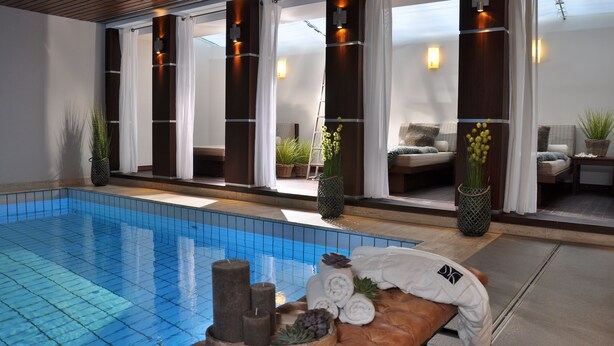 Uma área interna de piscina moderna com uma piscina azul cristalina, amplas janelas e um espaço de estar com sofás e plantas.