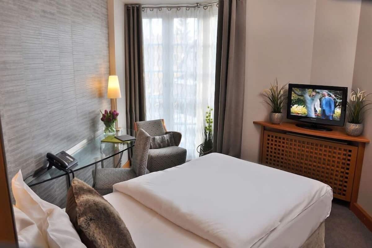 A hotel room with a bed, a desk with a telephone, a television, and a window with curtains.