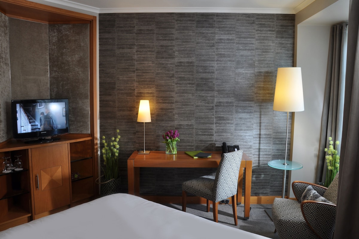 A hotel room with a bed, a desk with a lamp, a TV, and a chair.